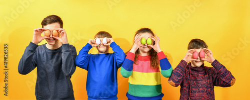 Four children hold Easter eggs to their faces instead of eyes on a yellow background.