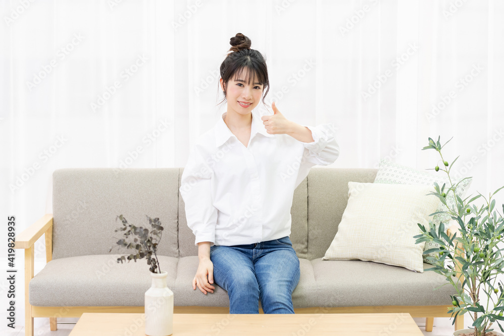窓際のソファに座って、いいねのジェスチャーをする若い女性