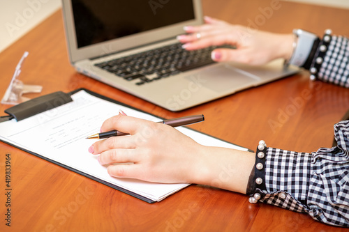 Female hands holding pen under the document and working with laptop at the table in an office