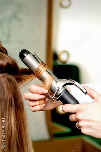 Back view of female hairdresser's hands curling women's hair with curling iron in a hair salon