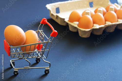 A supermarket cart filled with quality chicken eggs sits next to a carton of the same eggs. The concept of shopping in the store before Easter. Blue background photo