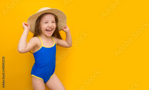 A beautiful cute little girl in a blue swimsuit laughs cheerfully and holds on to a straw hat. Mockup. Isolated yellow background. Summer vacation concept. Place for text