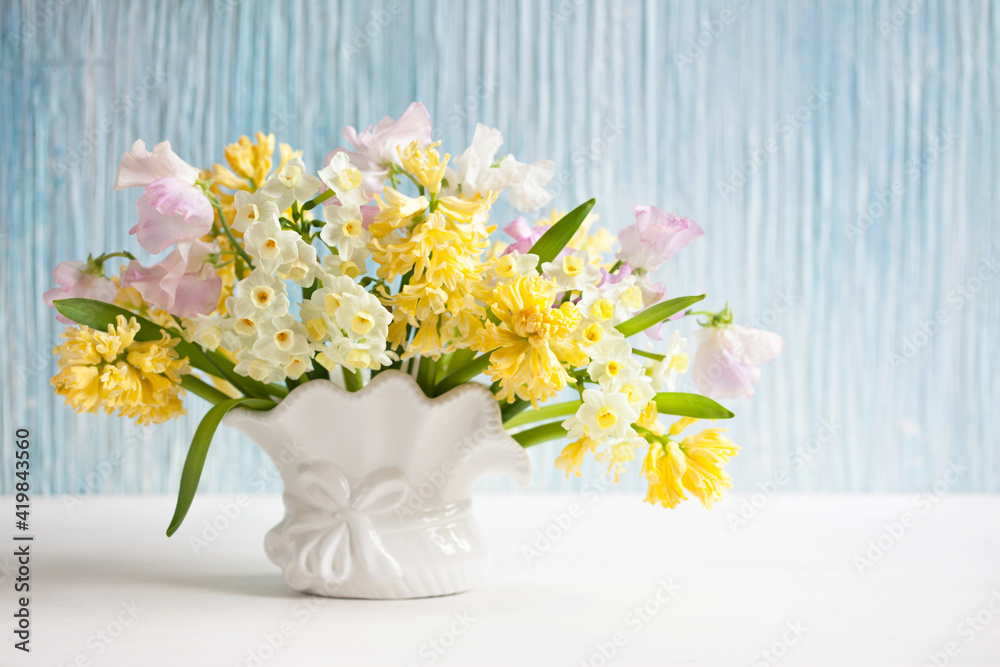 Spring bouquet of yellow hyacinths, daffodils and pink sweet peas in a vase on a table, blue background wall. Closeup, blur, soft focus.