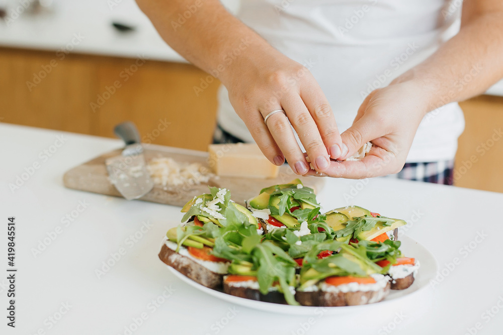 Young man making toasts with avocado, tomatoes, arugula, cheese in modern kitchen, putting parmesan