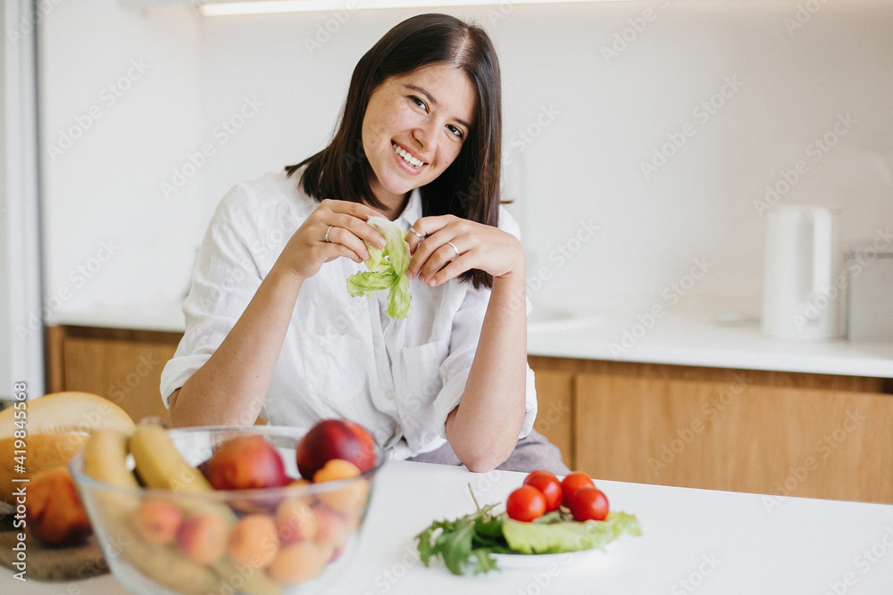 Young happy woman holding green lettuce leaf in hands and smiling in modern kitchen. Healthy eating