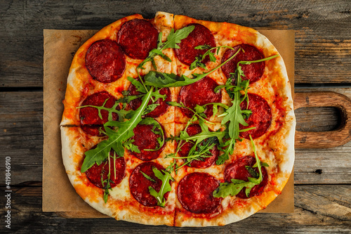 A pepperoni pizza with rocket salad on a wooden board, wooden rustic backdrop