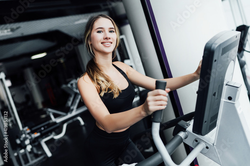 Attractive girl fitness trainer on cardio machine looks at camera and smiles