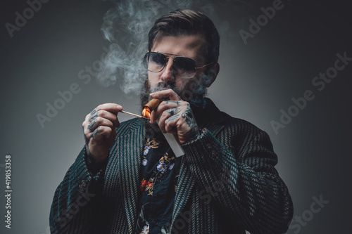 Portrait of a serious and handsome man with stylish coiffure posing in gray background. Tattooed man smoking cigar.