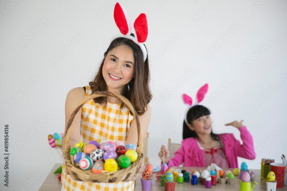 Beautiful asian woman wearing a bunny ears headband holding a selective focus of painted eggs. She feeling happy. In the background, her daughter painting the easter eggs with fun.