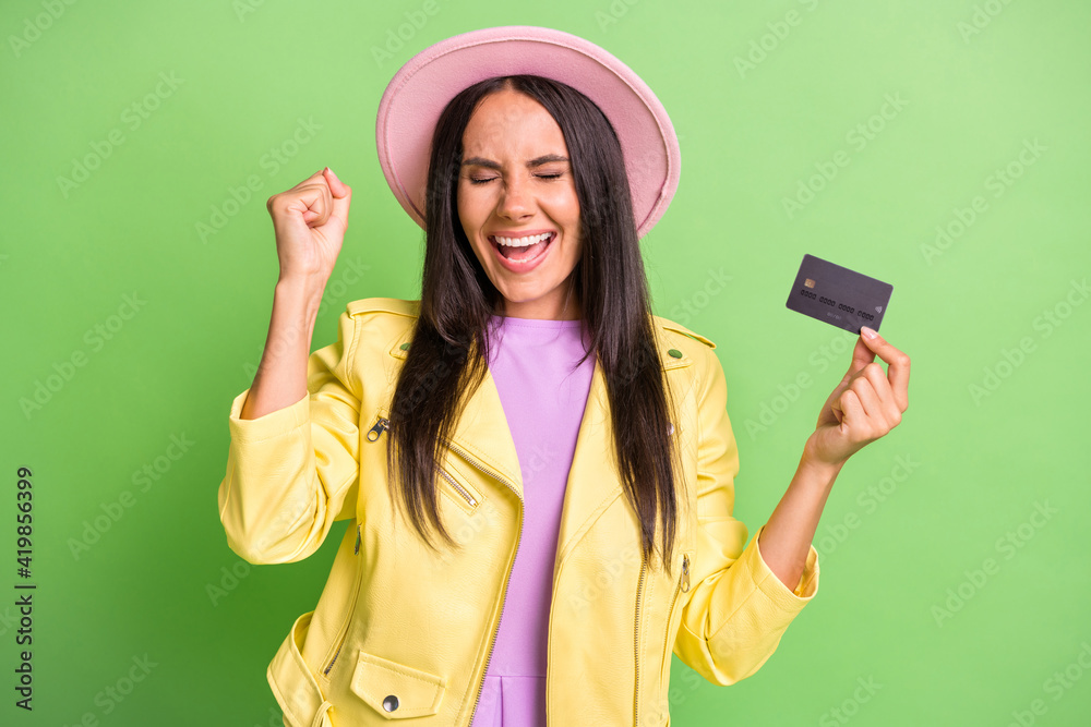 Photo of funky amazed girl hold credit card raise fist close eyes wear yellow jacket pink hat isolated green color background Photo of funky amazed girl hold credit card raise fist close eyes wear yellow jacket pink hat isolated green color background