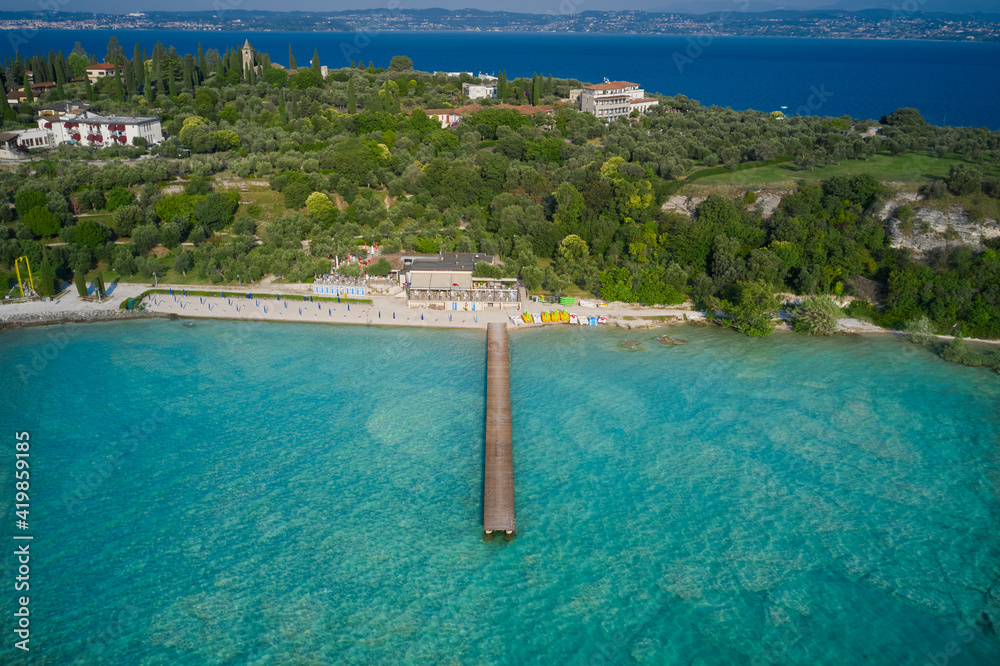 Wooden pier, turquoise water. Panoramic aerial view of Lido delle Bionde beach, Sirmione, Lake Garda, Italy. Island against the blue sky