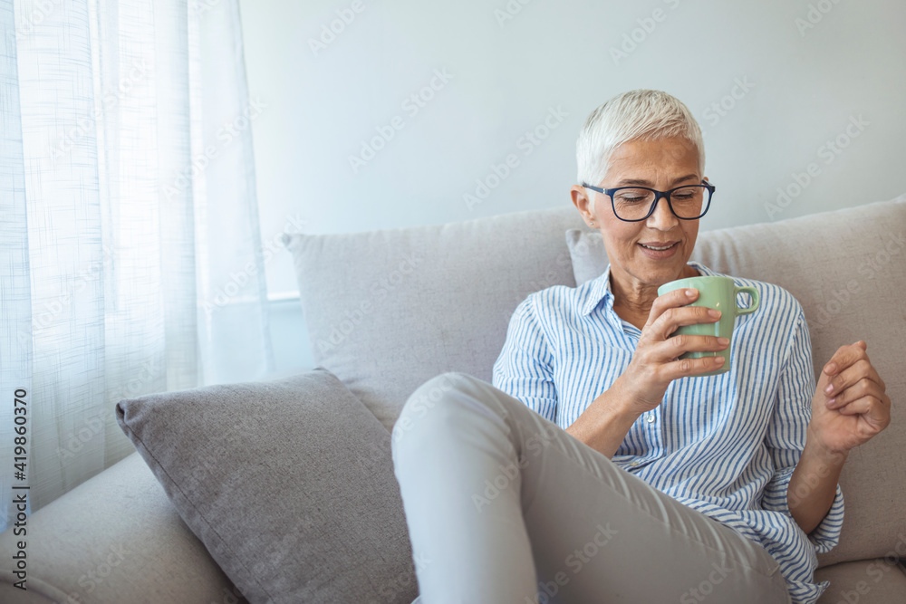 Happy beautiful relaxed mature older adult grey-haired woman drinking coffee relaxing on sofa at home. Smiling stylish middle aged 60s lady enjoying resting sitting on couch in modern living room. Happy beautiful relaxed mature older adult grey-haired woman drinking coffee relaxing on sofa at home. Smiling stylish middle aged 60s lady enjoying resting sitting on couch in modern living room.