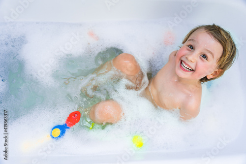 Little boy playing with toy by taking  bath in bathtub. Smiling toddler  sitting in bathtub with lot of fluffy soap bubble and smiling. Daily routine with children.