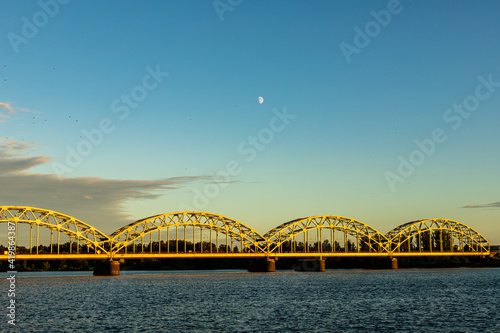 Riga´s railway Iron Bridge over Daugava River during sunset what makes it yellow, river water dark blue, half-moon is shining, black birds are flying in gradient blue sky 