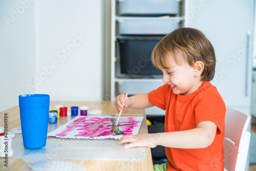 Little happy boy sitting at the desk and drawing colorful picture with paint and brushes. Child education at home during self isolation and lockdown. Concept of art and creativity of children.
