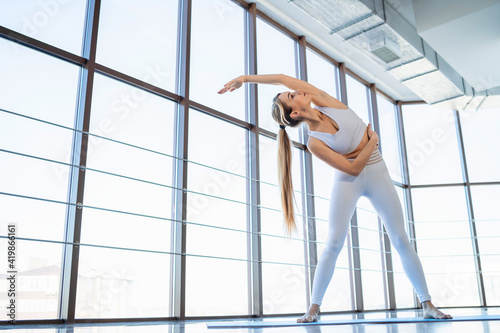 Beautiful girl in white sportswear warms up against the background of panoramic windows