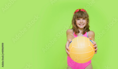 A beautiful child in a pink swimsuit shows a beach volleyball ball to the camera. Plortret of a cute little girl with swimming goggles on a green background. The concept of summer holidays.
