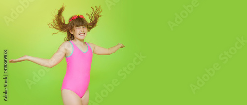 Banner of a beautiful pre-teenage girl in a pink swimsuit jumping cheerfully on a green background. Mockup. Funny child in a bathing suit with flying hair and arms to the side. Place for text