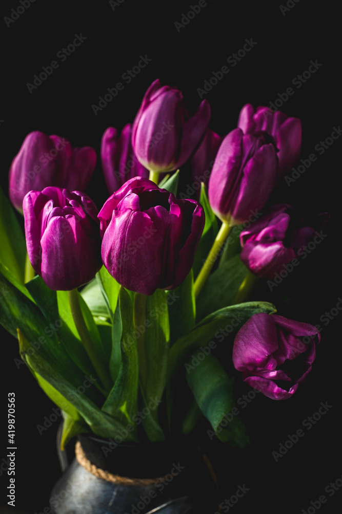 Fototapeta premium Lilac purple tulip flowers on table. Spring bouquet flowers in vintage vase. Floral concept. Floral background. Dark black background.