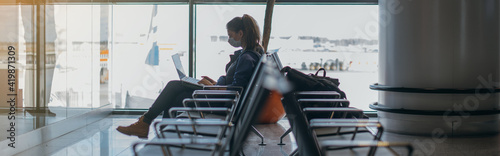 A woman is sitting at the airport with a laptop in a medical mask