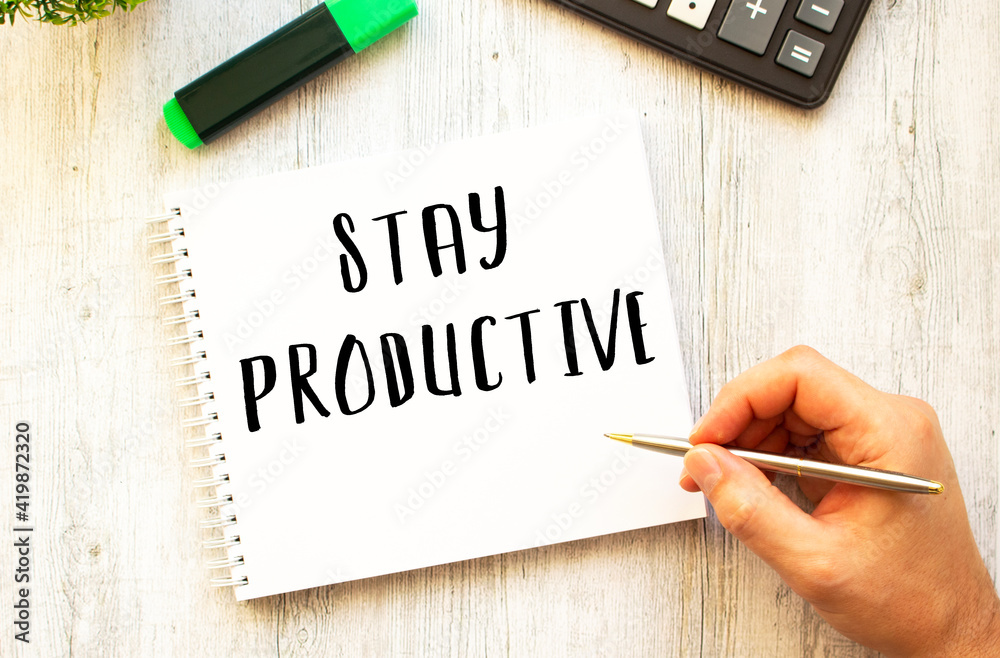 A hand with a pen writes text STAY PRODUCTIVE in a notebook on a wooden desk. View from above. A hand with a pen writes text STAY PRODUCTIVE in a notebook on a wooden desk. View from above.