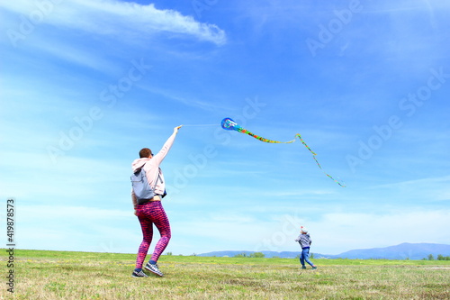 Happy family, mom and son, playing with flying kite on meadow in beautiful spring sunny day, blue sky in background