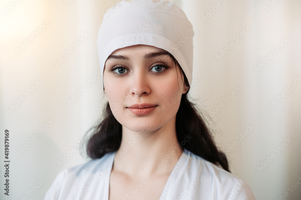 Nurse young medic standing near white wall and looking at the camera. Female doctor dressed in medical gown and surgical hat. Image with copy space. Pandemic virus coronavirus concept Nurse young medic standing near white wall and looking at the camera. Female doctor dressed in medical gown and surgical hat. Image with copy space. Pandemic virus coronavirus concept