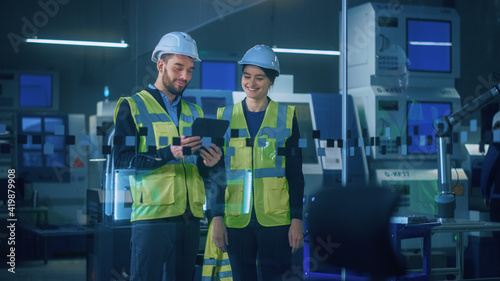 Modern Factory: Female and Male Engineers Wearing Safety Jackets, Hardhats Standing in Industrial Workshop, Talking and Using Tablet Computer. Facility with CNC Machinery, Robot Arm Production Line