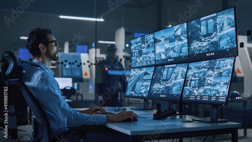 Industry 4.0 Modern Factory: Security Operator Controls Proper Functioning of Workshop Production Line, Uses Computer with Screens Showing Surveillance Camera Feed. High-Tech Security