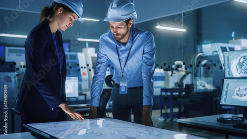 Industry 4.0 Modern Factory Office Meeting: Female Manager and Male Engineer Talking, Using Touchscreen Digital Table to Inspect and Analyze Machinery Blueprints. High-Tech Electronics Facility 