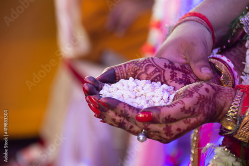 The bride's brother puts puffed rice in the hands of the bride, and the groom standing close to her holds her hands from the back. They then pour the offering into the fire together.