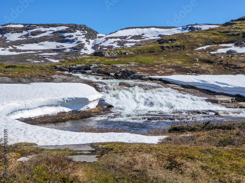 A wast river flowing in higher parts of Eidfjord, Norway. Some parts of the river are covered with a snow layer. Nature wakes up after winter. Lush green flora growing on the slopes. Sky is clear blue