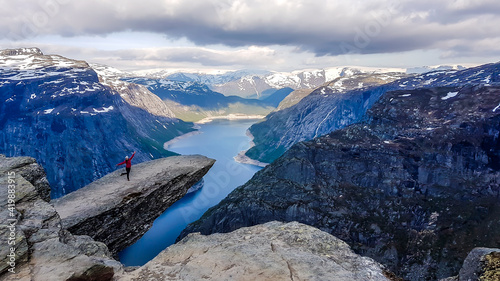 A girl wearing pink jacket stands at the hanging rock formation, Trolltunga with a view on Ringedalsvatnet lake, Norway. Slopes of the mountains are partially covered with snow. Freedom and happiness