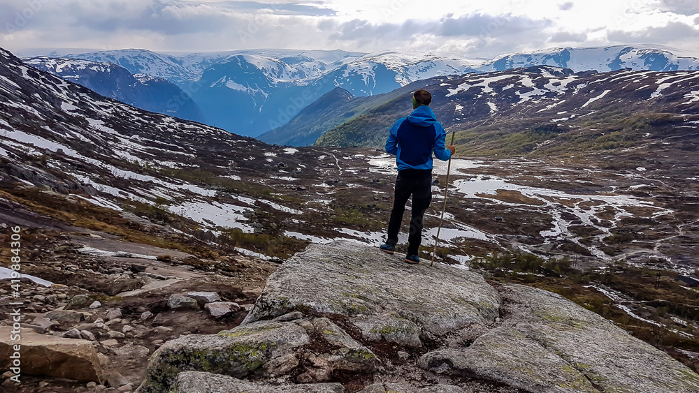 A young man wearing blue jacket stands on a pile of rocks and admires the view in front of him. He supports himself on a wooden stick. Endless mountain ranges partially covered with snow. Big overcast