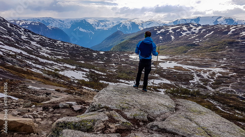 A young man wearing blue jacket stands on a pile of rocks and admires the view in front of him. He supports himself on a wooden stick. Endless mountain ranges partially covered with snow. Big overcast