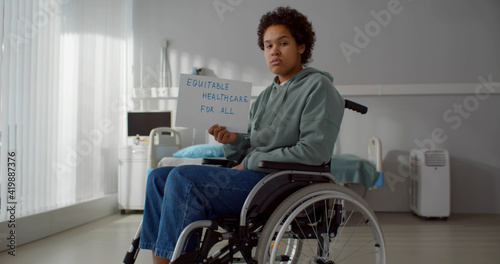 Upset afro patient in wheelchair at hospital ward and holding equitable health care for all poster