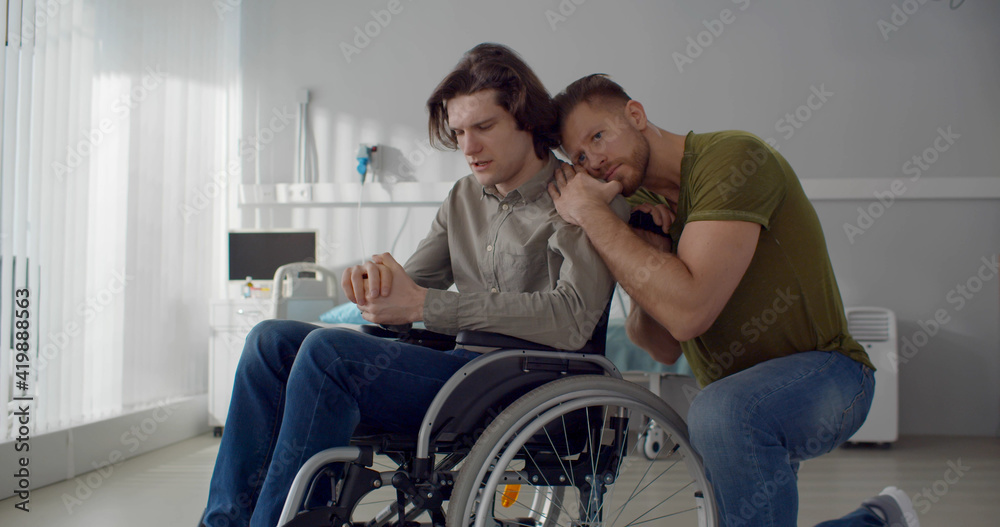 Young man hugging and supporting depressed boyfriend in wheelchair