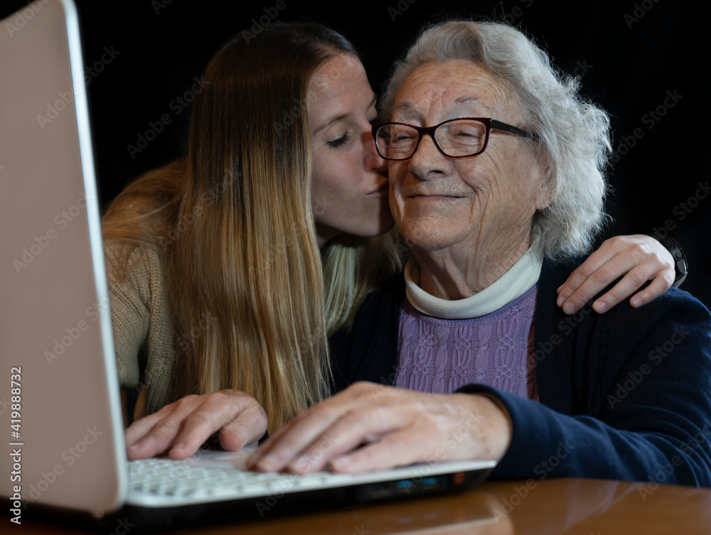 Young granddaughter giving a hug and kissing old 80s woman grandma while is teaching to use laptop at home.Family spending time and having fun together with new technologies concept lifestyle. Young granddaughter giving a hug and kissing old 80s woman grandma while is teaching to use laptop at home.Family spending time and having fun together with new technologies concept lifestyle.