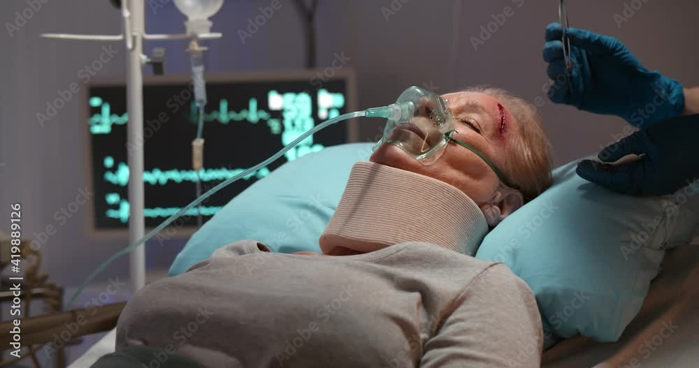 Injured woman with neck brace and oxygen mask in hospital bed, nurse treating forehead wound