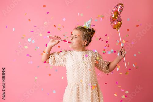 A happy little girl in a birthday cap holds a doughnut-shaped balloon on a pink background in the studio, space for text
