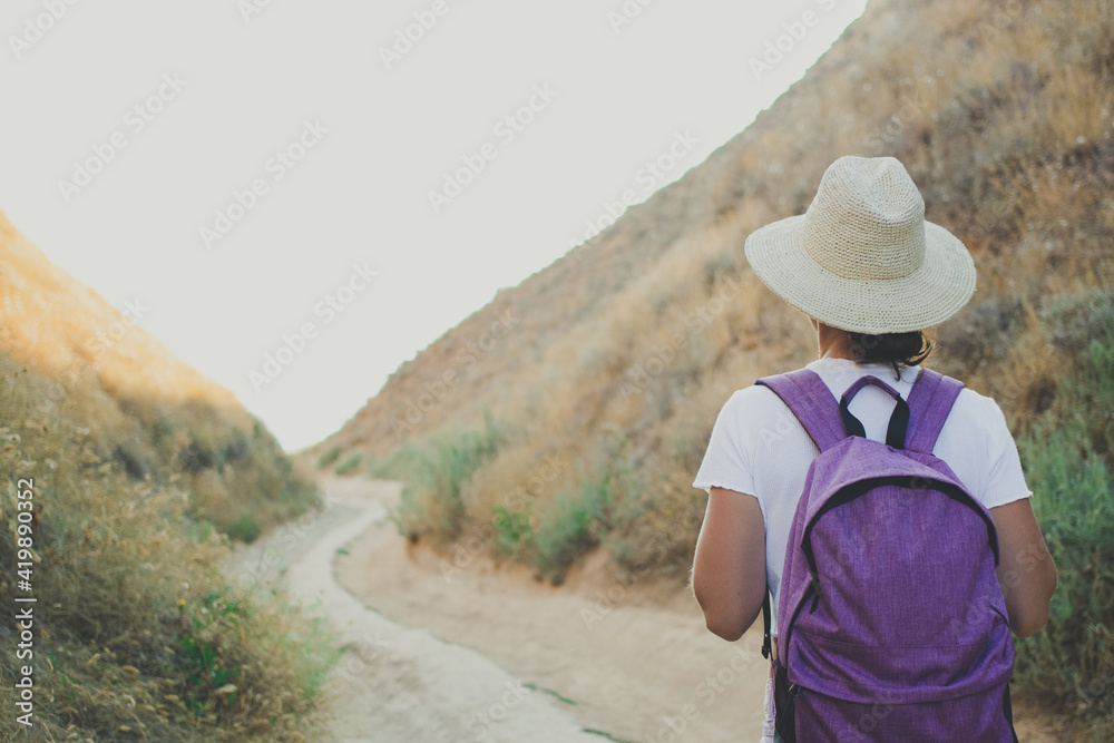 Back view of woman hiker traveler walking on path in canyon gorge of national landscape park,wearing purple backpack, summer straw hat and white t-shirt.Active travel and adventure concept.Copy space