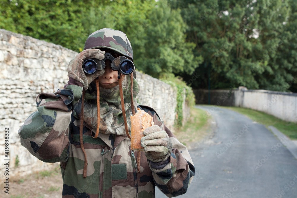 A masked boy ready to go hunting holds binoculars and a sandwich