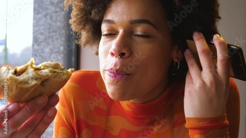 Young beautiful multiethnic woman indoors restaurant biting hamburger using smartphone - hunger, delicious, eating concept