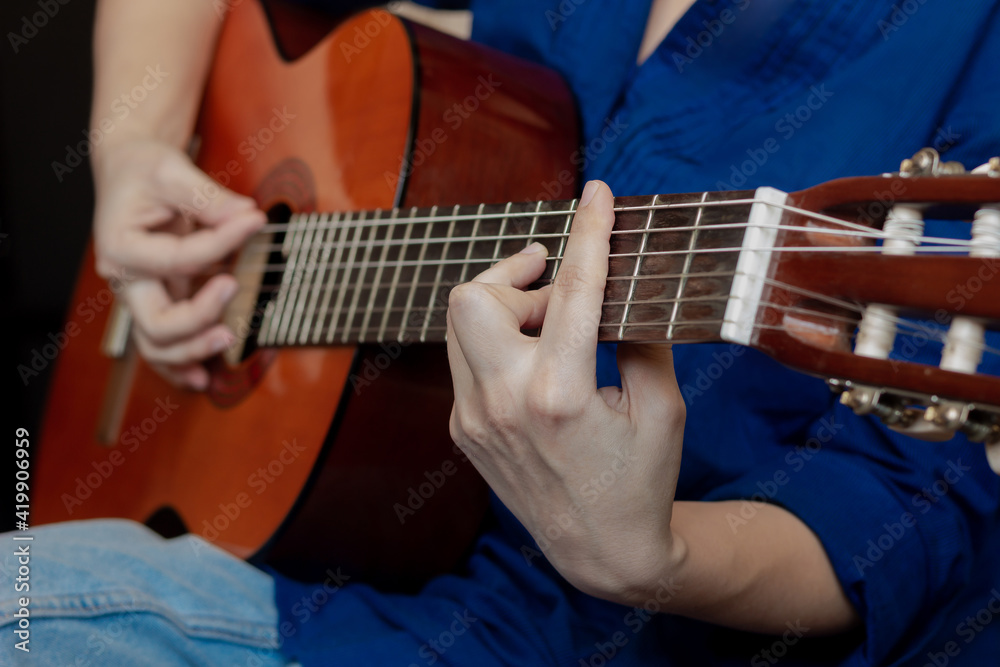 Fototapeta premium Close up of hand of young woman in the blue shirt and jeans playing acoustic guitar. Girl picks a barre chord clamping frets on the fretboard