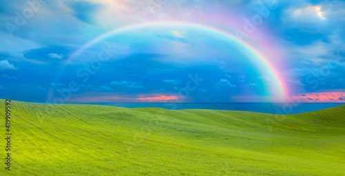 Green grass field with rainbow at sunset, green sea in the background
