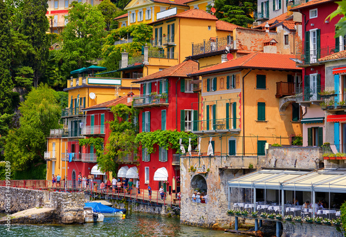 Varenna, Italy. Picturesque town at lake Como. Colourful motley Mediterranean houses at stone beach coastline among green trees. Popular health resort and touristic location. Summer day landscape.