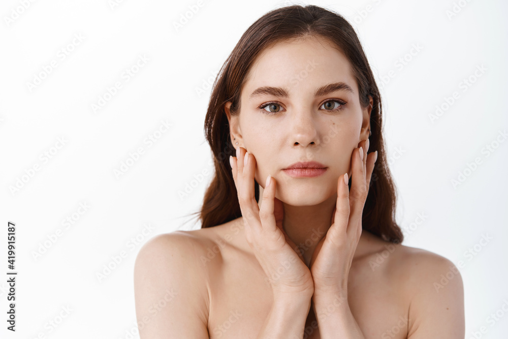 Skin care. Young beauty woman with natural make up, touching hydrated and nourished facial skin, holding hands on cheeks, standing naked against white background