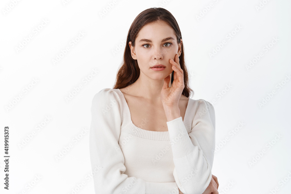 Skin care and cosmetics. Young woman with nude natural make up, touching hydrated clean facial skin, holding hand on cheek, gazing at camera thoughtful, white background Skin care and cosmetics. Young woman with nude natural make up, touching hydrated clean facial skin, holding hand on cheek, gazing at camera thoughtful, white background