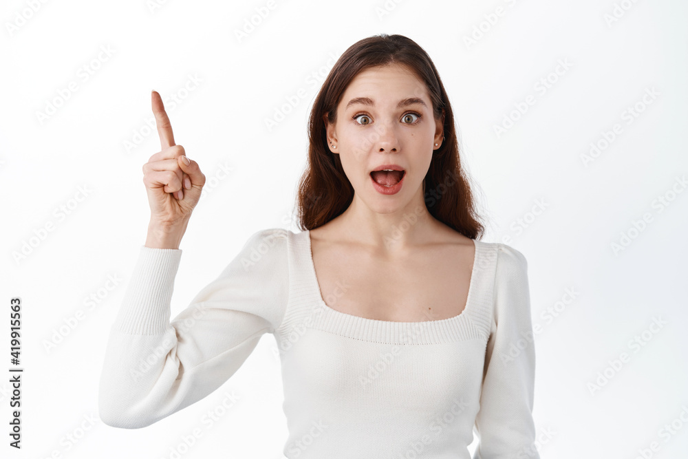 Check this out. Surprised and excited brunette girl showing awesome news, pointing finger at upper left corner, showing logo on copy space aside, standing in studio on white background Check this out. Surprised and excited brunette girl showing awesome news, pointing finger at upper left corner, showing logo on copy space aside, standing in studio on white background