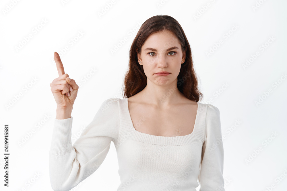 Disappointed and sad girl pointing at unfair bad thing, points up at logo on top and pouting upset, standing displeased against white background Disappointed and sad girl pointing at unfair bad thing, points up at logo on top and pouting upset, standing displeased against white background